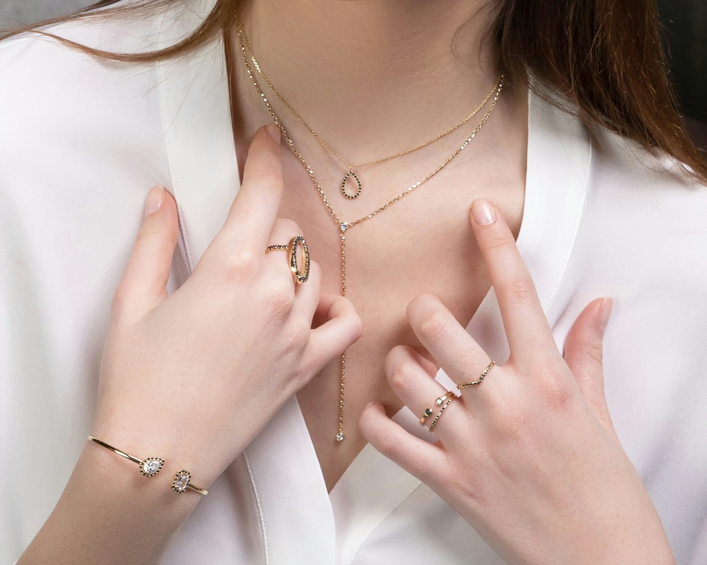 Hands resting at the neckline wearing layered gold necklaces, rings and a fine bracelet
