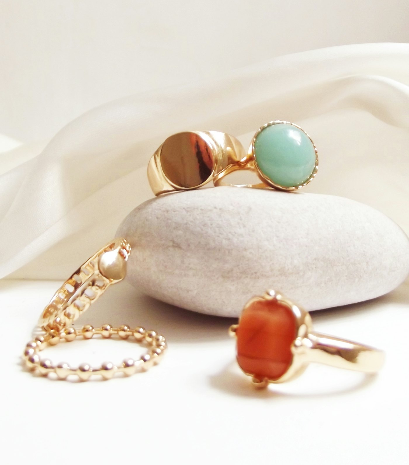 Gold rings set with orange and green stones arranged on a smooth cream pebble